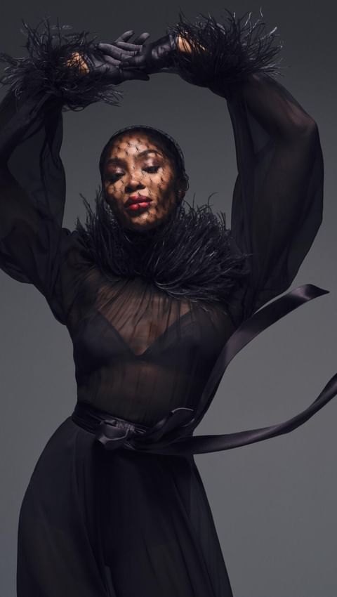 An elegant woman in a flowing black dress with feathered accents, captured in a studio setting with a high-fashion editorial aesthetic.