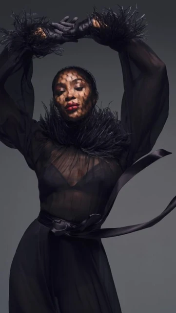 An elegant woman in a flowing black dress with feathered accents, captured in a studio setting with a high-fashion editorial aesthetic.