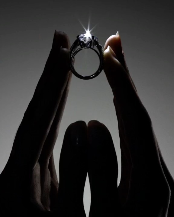 Close-up of a woman's hands holding up a diamond ring against a dark background, highlighting its sparkle and design.