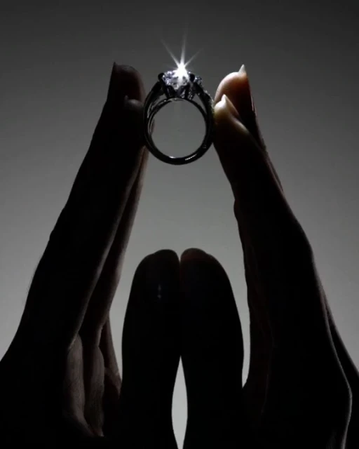 Close-up of a woman's hands holding up a diamond ring against a dark background, highlighting its sparkle and design.