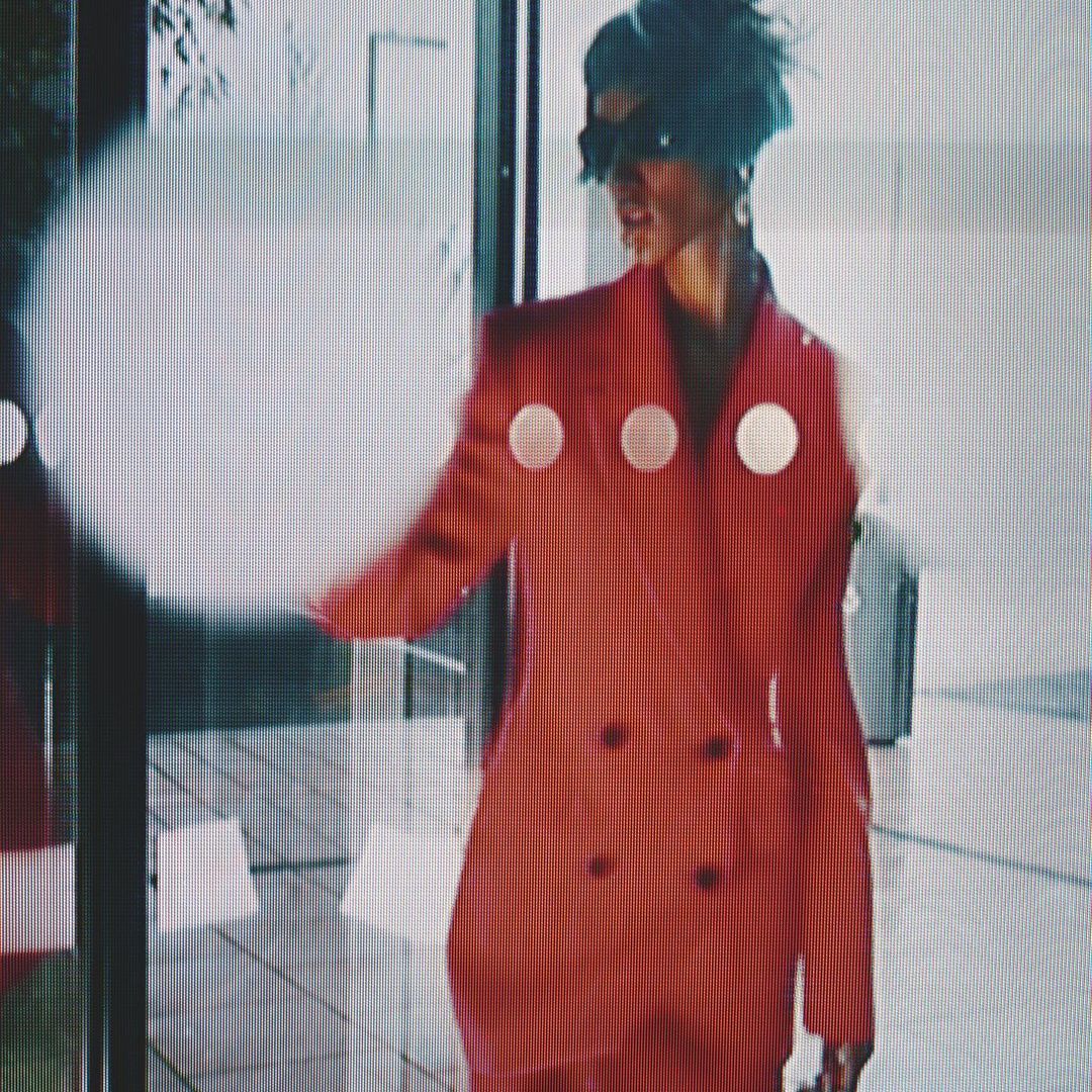 A woman in a red blazer with white circles stands in front of a building with a glass window and door, creating a modern, stylish scene.