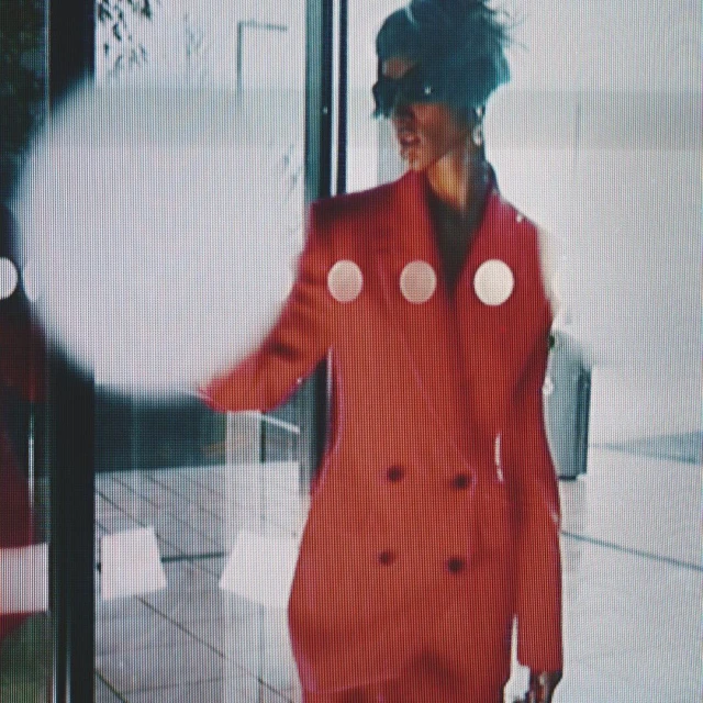 A woman in a red blazer with white circles stands in front of a building with a glass window and door, creating a modern, stylish scene.