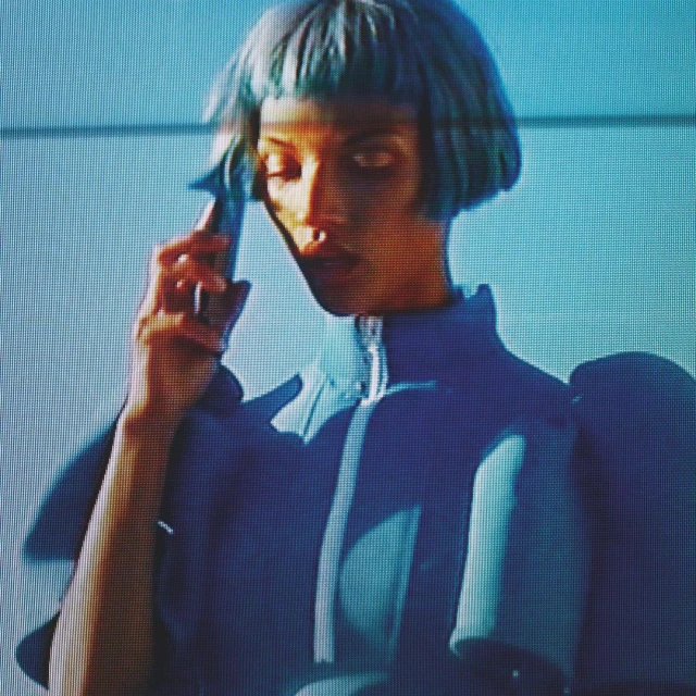 Woman with blue hair and a futuristic outfit stands in front of a blue wall. Fashion forward portrait with unique style.