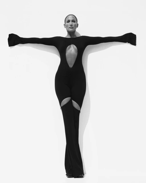 Monochrome portrait of Jennifer Lopez posing in a black cutout bodysuit against a white backdrop, arms outstretched and legs apart.