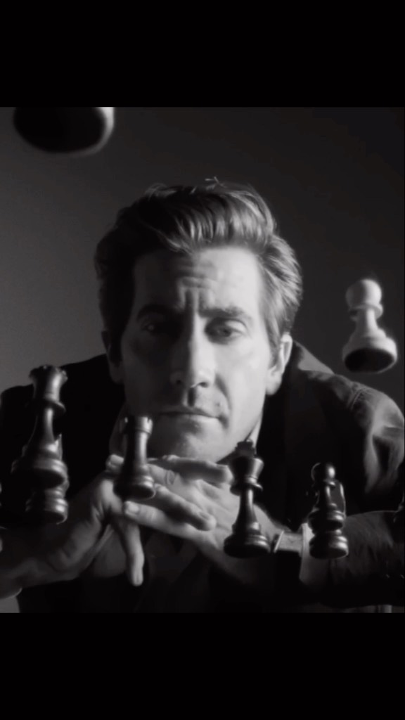 Black and white portrait of an adult male actor gazing forward surrounded by floating chess pieces in an indoor studio environment.