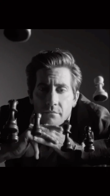 Black and white portrait of an adult male actor gazing forward surrounded by floating chess pieces in an indoor studio environment.