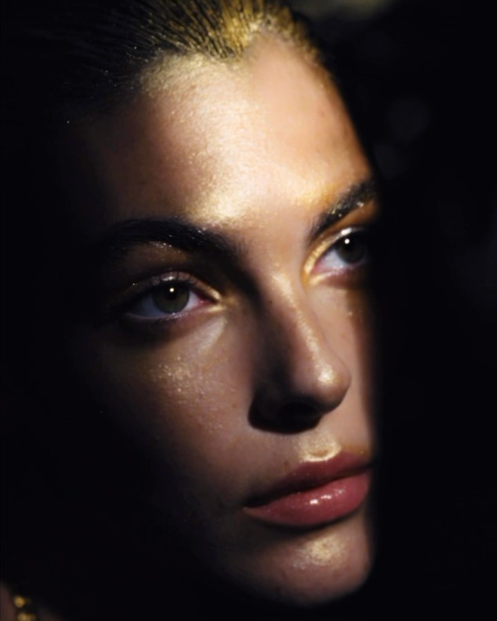 Close-up portrait of an adult female with shiny, golden makeup and dark background, looking upward in a dimly lit setting, beauty shot.