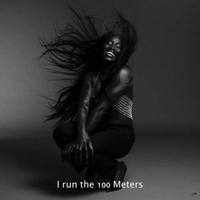 Striking monochrome portrait of a smiling Black woman with flowing hair, wearing a studded top and dark pants, captured in a studio setting.