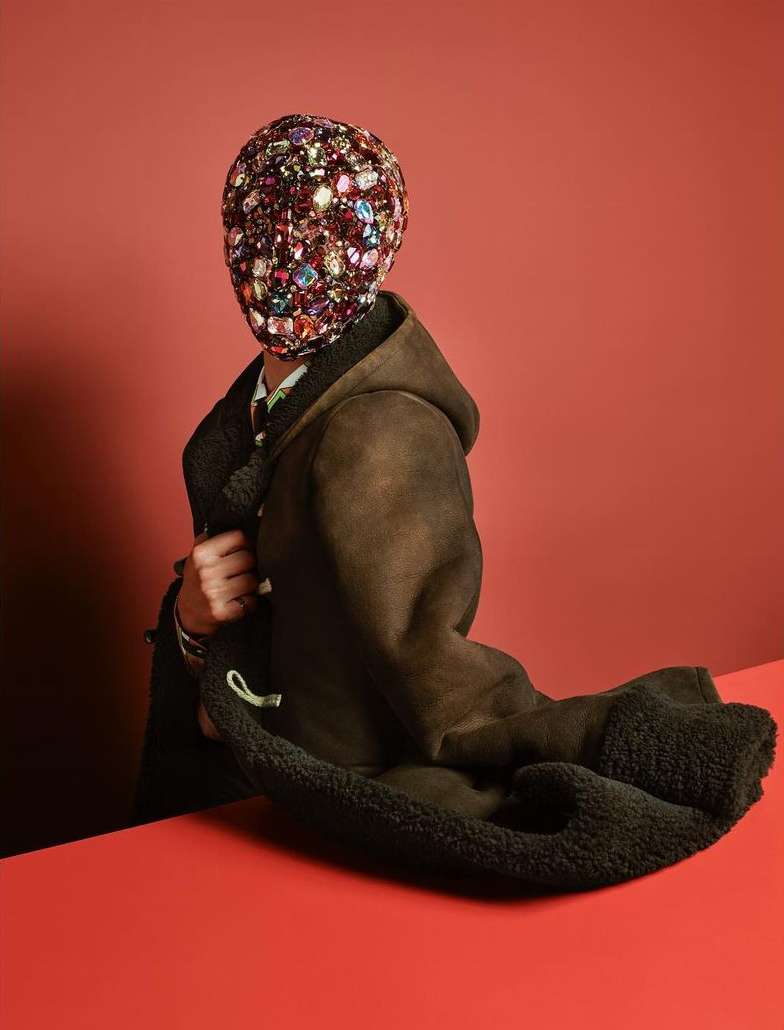 A person is standing in a photo studio wearing a long, brown coat with a colorful head covering and standing in front of a red background.