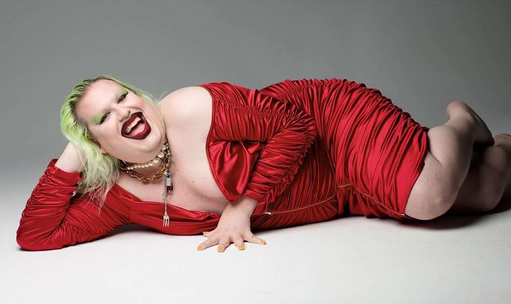 Person in red dress lying down with bright green hair and painted yellow fingernails laughing in a studio setting with a light gray backdrop.