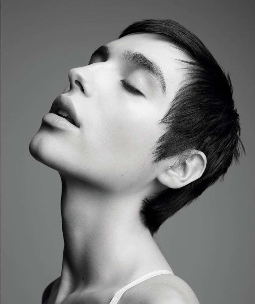 Monochrome portrait of a young person with dark hair, closed eyes, and light skin, set against a simple grey background in a photo studio.
