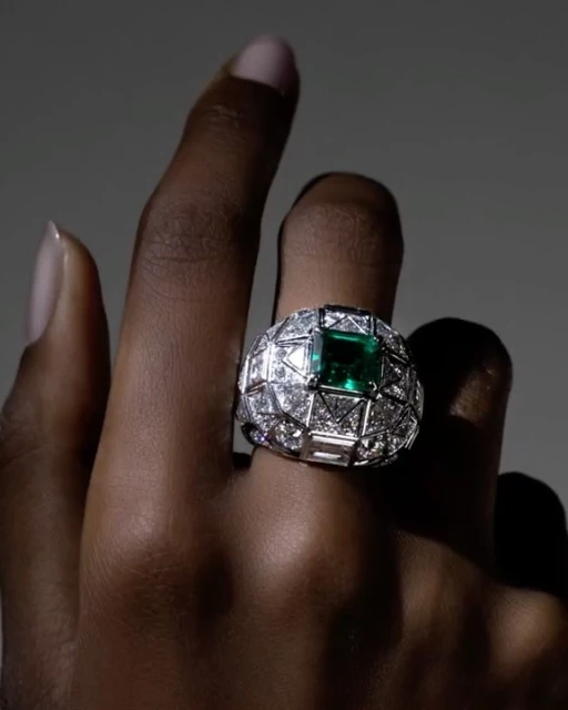 Close-up of a hand wearing a large, ornate ring with diamonds and a green emerald gemstone, under soft lighting, against a grey backdrop.
