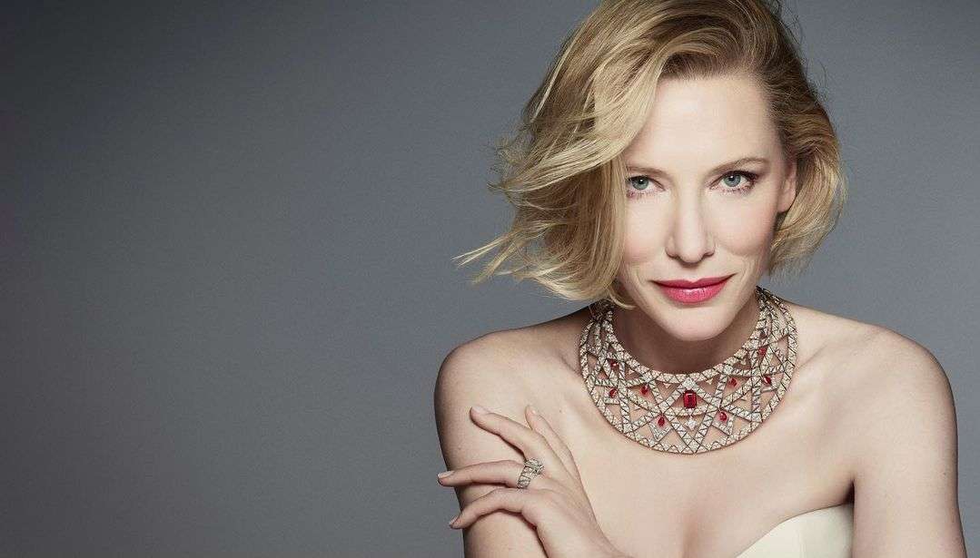 Close-up portrait of a blond woman with fair skin wearing an elaborate statement necklace against a solid gray background.