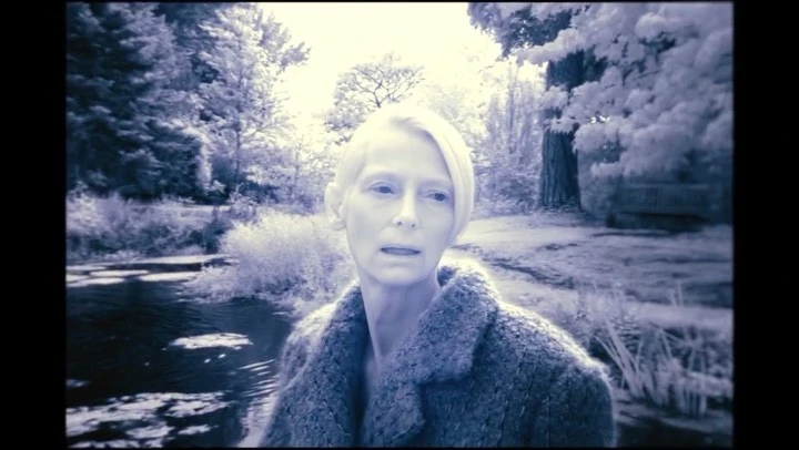 Monochrome shot of a fair-skinned woman in a coat standing in a forest landscape with water and trees under soft lighting.