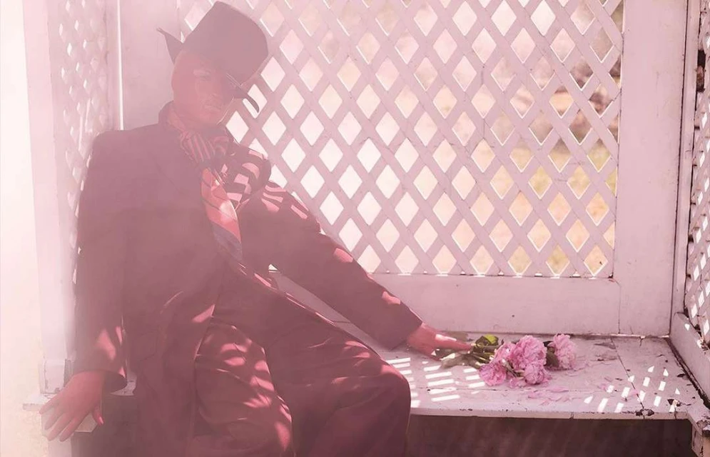 Person in a suit and hat sits on a bench with flowers in a lattice-walled gazebo, bathed in a soft pink light, creating a vintage aesthetic.