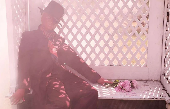 Person in a suit and hat sits on a bench with flowers in a lattice-walled gazebo, bathed in a soft pink light, creating a vintage aesthetic.