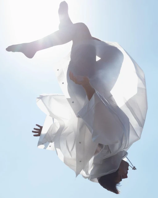 A woman in mid-air, upside down, wearing a white coat, set against a clear blue sky. Her limbs are extended, creating a sense of motion and freedom.