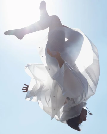 A woman in mid-air, upside down, wearing a white coat, set against a clear blue sky. Her limbs are extended, creating a sense of motion and freedom.