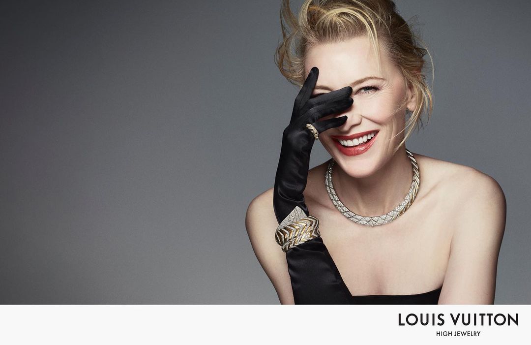 Smiling woman with blond hair wearing a necklace and bracelet, partially covering her face with a gloved hand against a gray background.