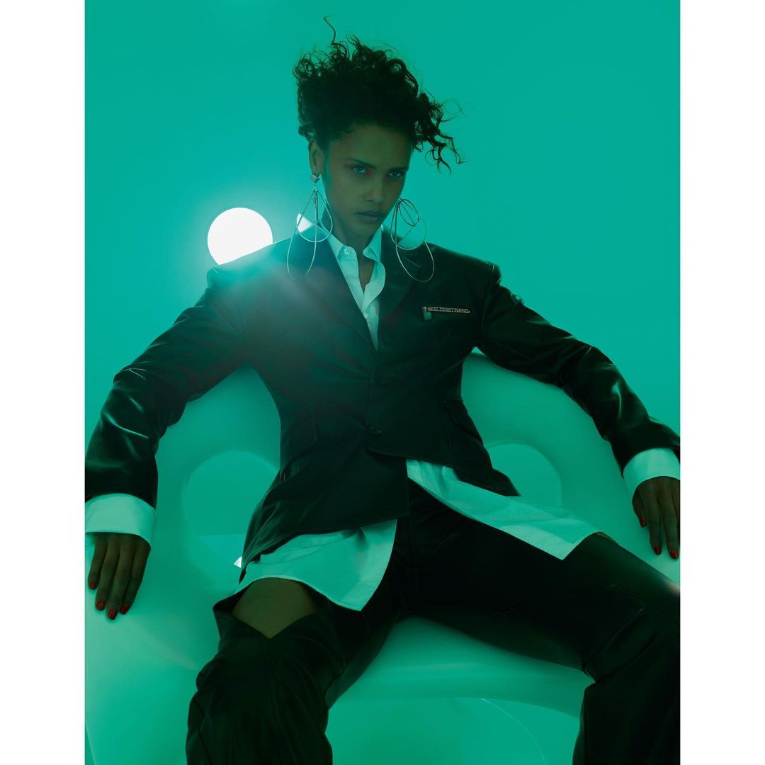 Fashionable woman in blazer and thigh-high boots poses in a studio against a teal background with a bright light source.