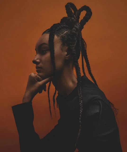 Portrait of a woman with braided hair pulled up on top of her head, wearing a black top against an orange background.