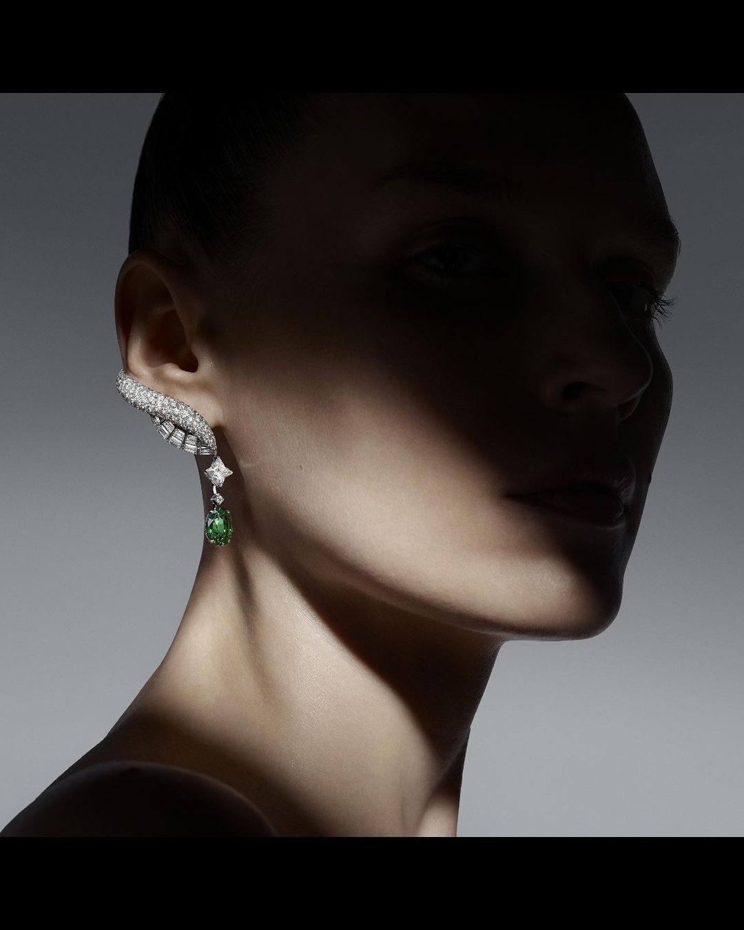 Close-up of a woman with a distinctive diamond and green gemstone earring, set against a gradient gray background with dramatic lighting.