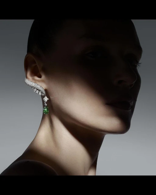 Close-up of a woman with a distinctive diamond and green gemstone earring, set against a gradient gray background with dramatic lighting.