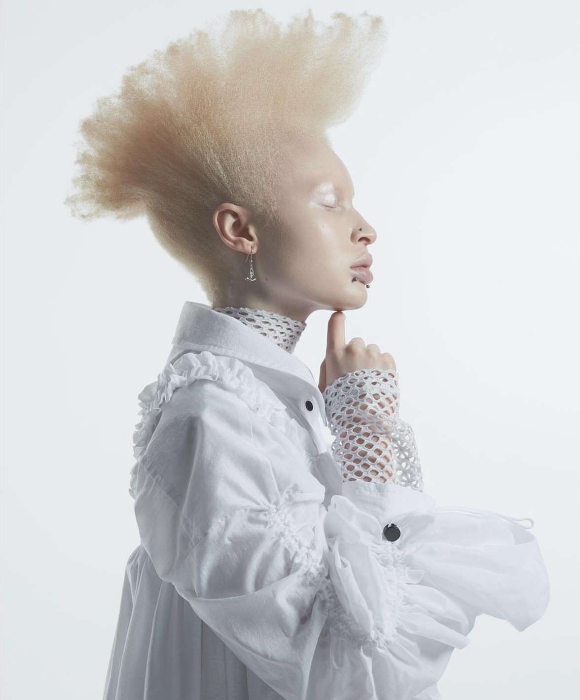 Close-up portrait of an adult albino woman with a high-top hairstyle, wearing a white ruffled top, against a bright white background.