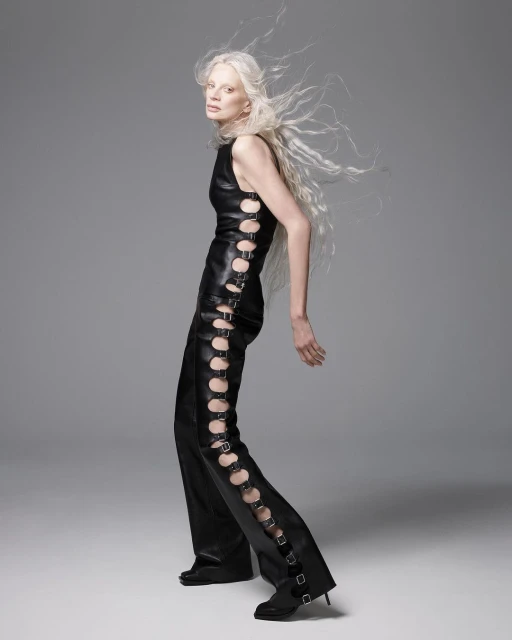 Adult woman in black leather sleeveless top and pants with side cut-outs poses in a studio with grey background and neutral lighting.