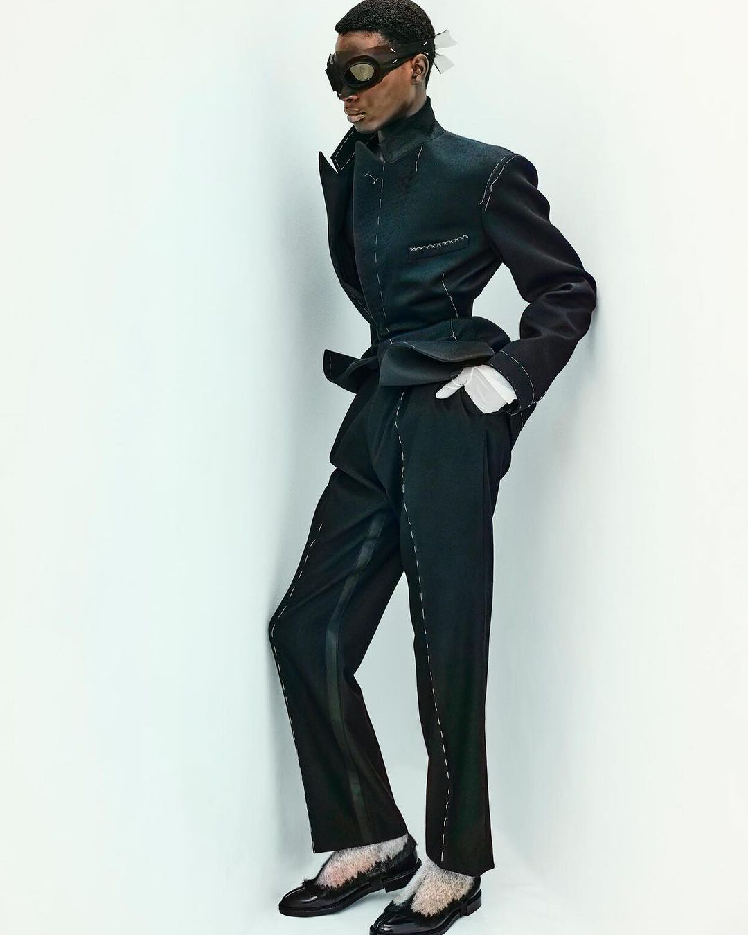 Fashion shot of a Black male model in a black suit with white stitching, posing in front of a plain white wall in a studio setting.