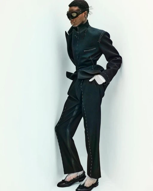 Fashion shot of a Black male model in a black suit with white stitching, posing in front of a plain white wall in a studio setting.