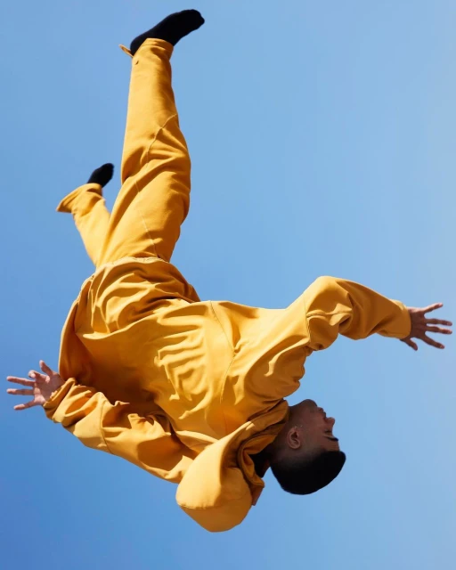 Low-angle shot of a man in a mustard-yellow jumpsuit falling against a clear blue sky, showcasing a dynamic and gravity-defying moment.