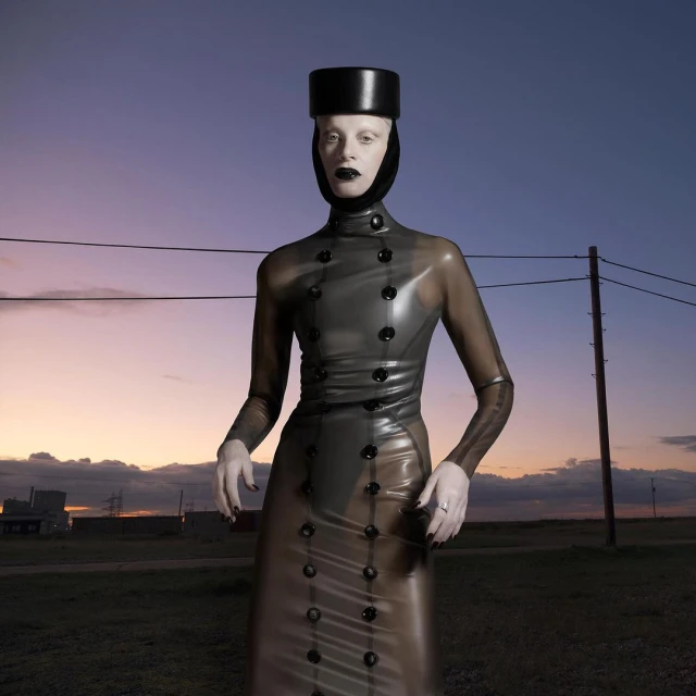 Woman in a translucent grey coat, hat, and dark makeup stands in a desolate landscape against a sunset sky and distant industrial buildings.