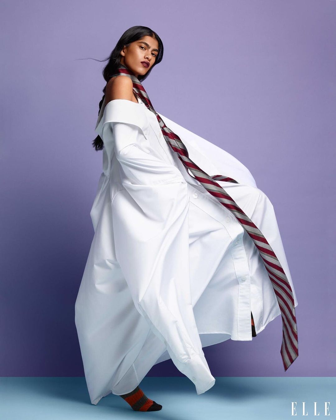 Fashion editorial shot of a woman in an oversized white shirt, tie, and sock, posing against a purple backdrop in a studio setting.