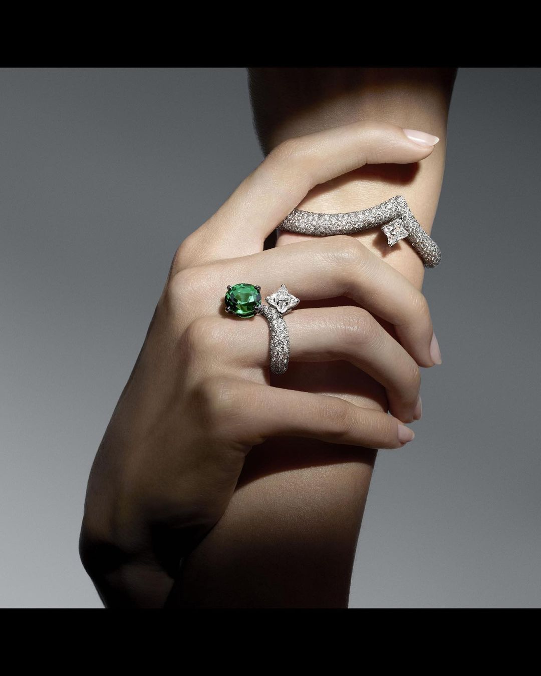 Closeup of fair-skinned hand with emerald and diamond rings, and diamond bracelet against soft grey background in beauty shot.