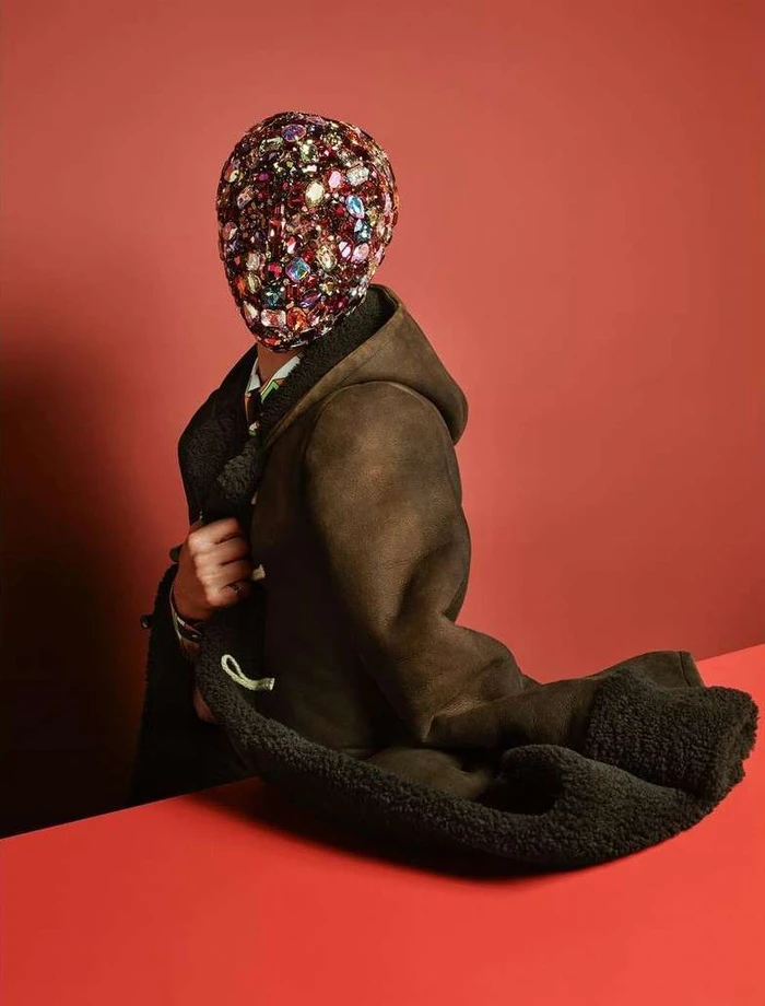 Model wearing a jewel-encrusted mask and a dark shearling coat against a warm coral background by Sølve Sundsbø