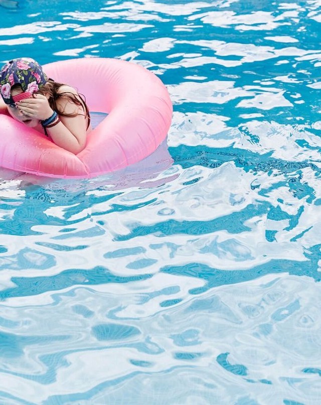 Child floats in swimming pool with pink inflatable ring, wearing a floral swim cap and covering her eyes with her hands.