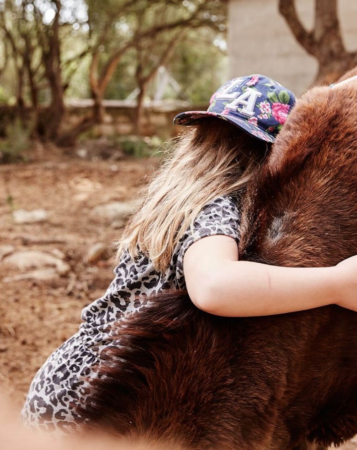 Girl in cap hugs brown furry animal outdoors in a rural setting with trees, rocks, and natural light, creating a tender scene.