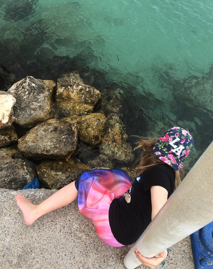 Child in floral hat sits on concrete near rocks and turquoise ocean water. The girl wears a dark shirt and colorful skirt.