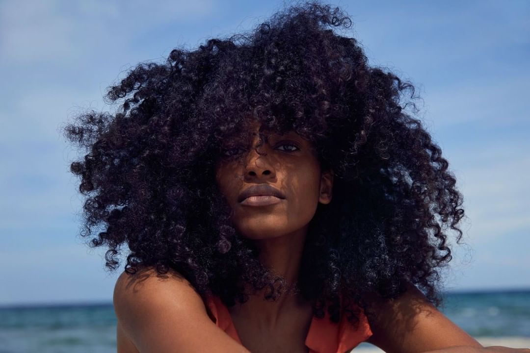 Close-up portrait of an adult woman with curly hair posing against a beach and sky background on a sunny day, focusing on natural beauty.