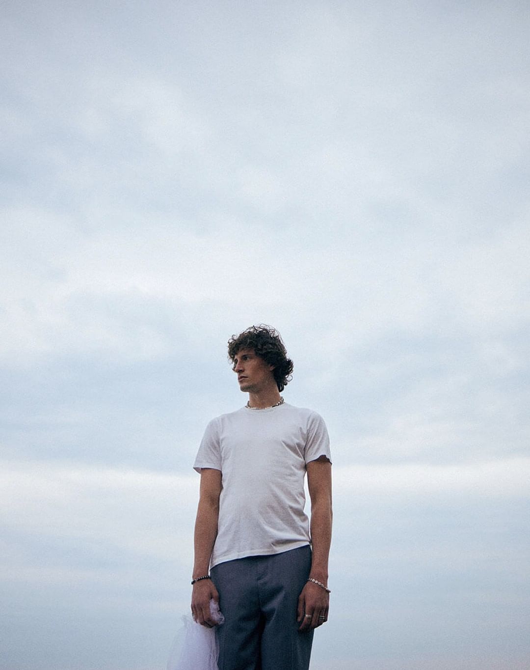 Young adult with curly hair stands outdoors against cloudy sky, wearing a white t-shirt and grey pants, holding white clothing.