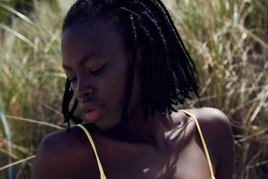 Close-up portrait of a Black woman with braided hair wearing a yellow strap top outdoors near tall grass.