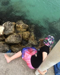 Young person sitting barefoot on a concrete pier next to rocks and turquoise water, wearing a floral cap and colourful skirt.
