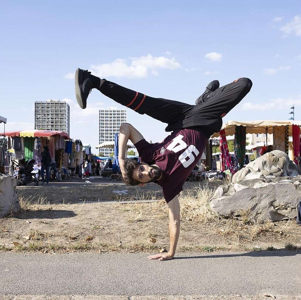 A man is doing a one-handed handstand outdoors in front of a clothing market and some apartment buildings under a partly cloudy sky.