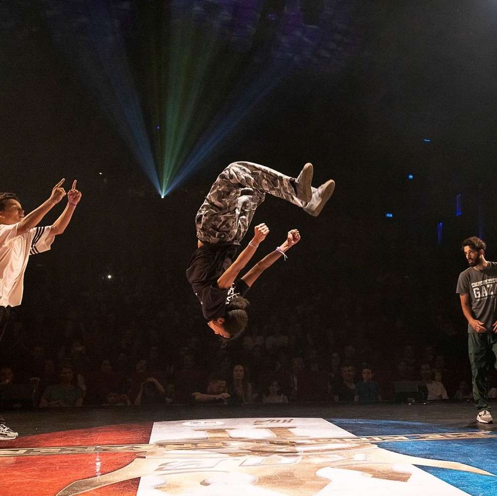 A young male breakdancer performs a backflip on a stage in front of a large crowd, illuminated by spotlights. Two other young men are on stage.