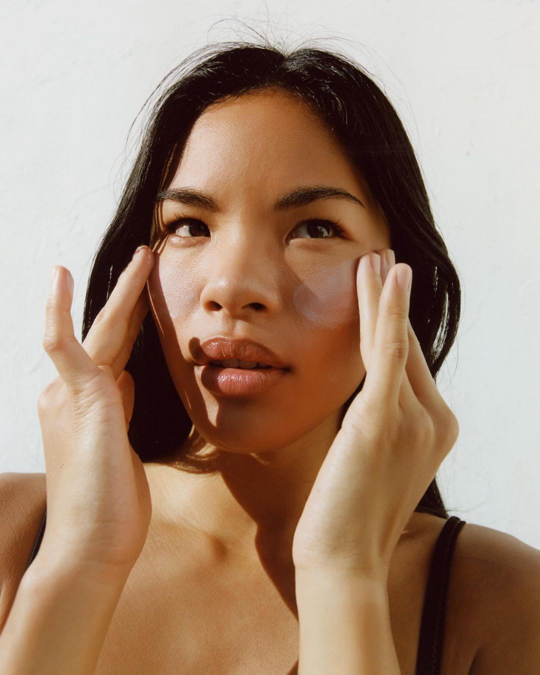 Close-up shot of a young adult woman with long, dark hair, applying skincare around her eyes in a bright, naturally lit indoor setting.
