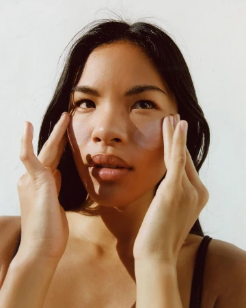 Close-up shot of a young adult woman with long, dark hair, applying skincare around her eyes in a bright, naturally lit indoor setting.