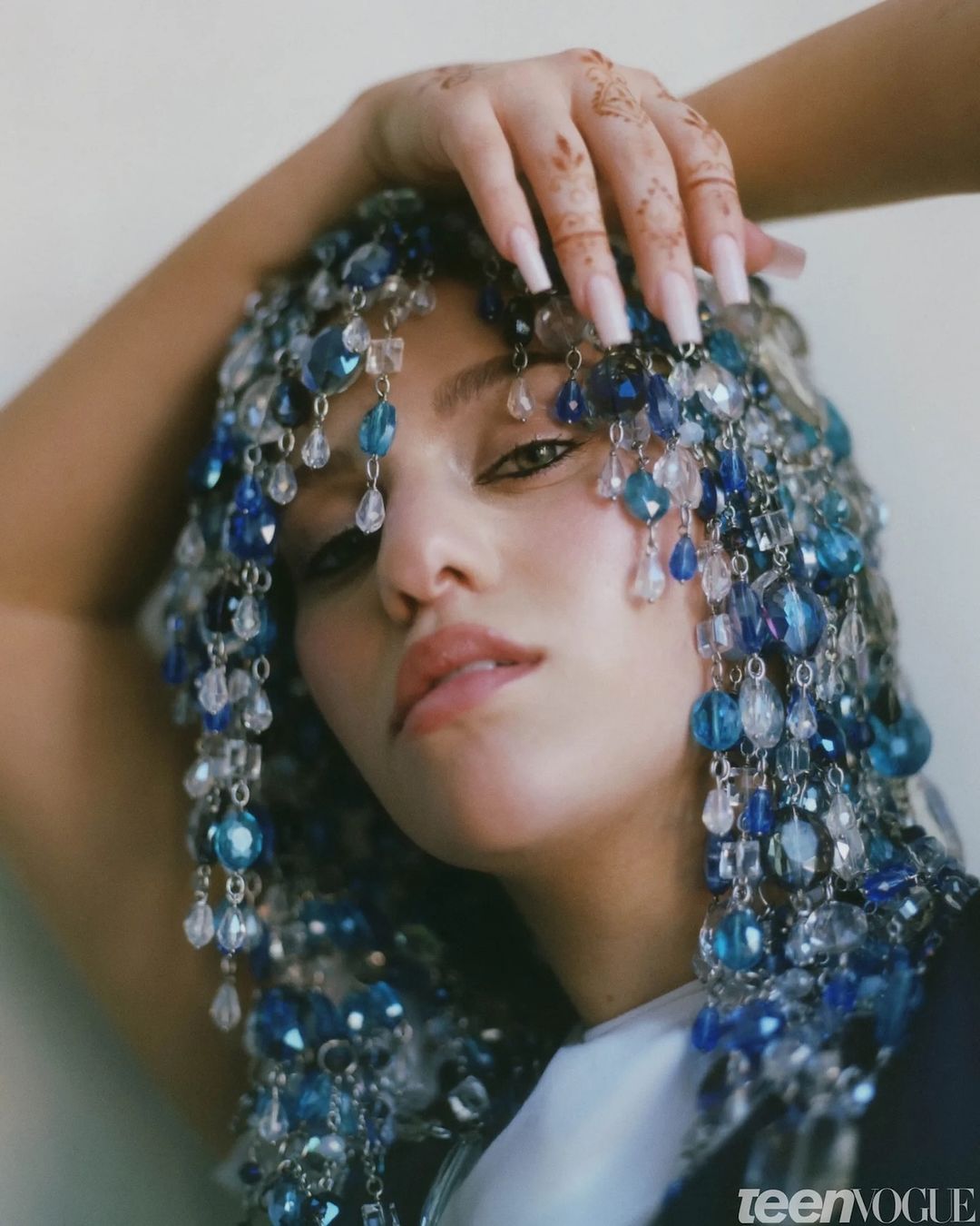 Close-up of a young adult woman posing with beaded headwear, hands on her head, and long nails, on a neutral background.