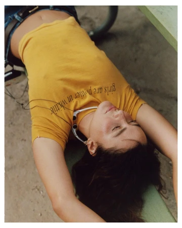Woman in yellow t-shirt lies on a green surface outdoors, eyes closed, wearing headphones, jeans and a belt, with dark hair.