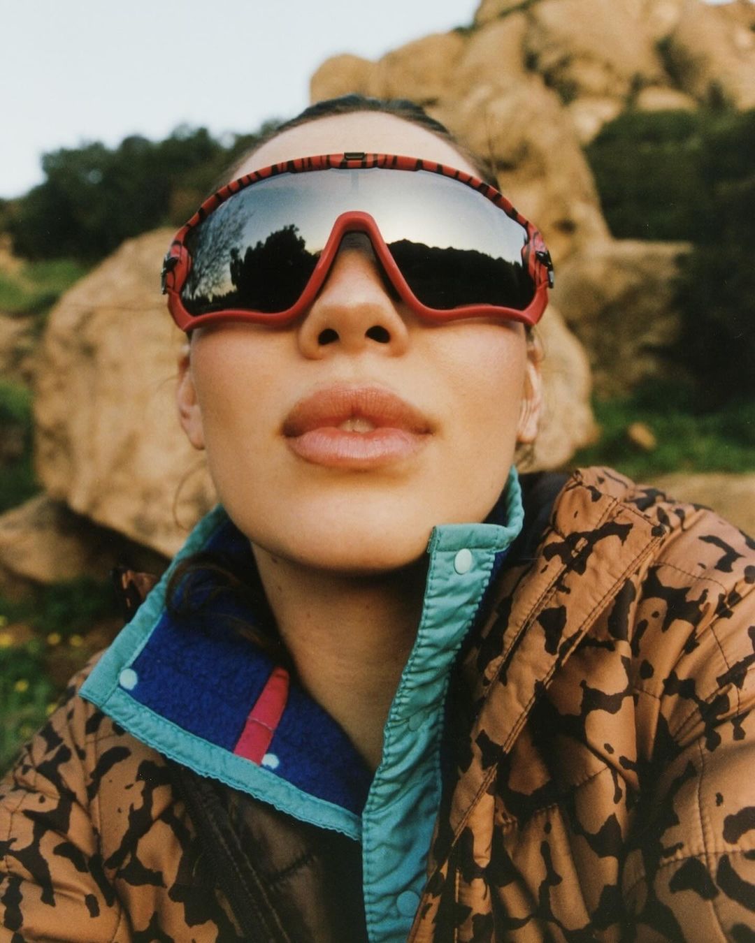 Outdoor portrait of a woman wearing sporty sunglasses and jacket with a rocky hill landscape and trees in the background.